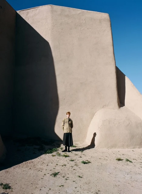 A lone woman stands in front of an adobe wall with shadows under a clear blue sky, creating a stark and minimalist outdoor scene.