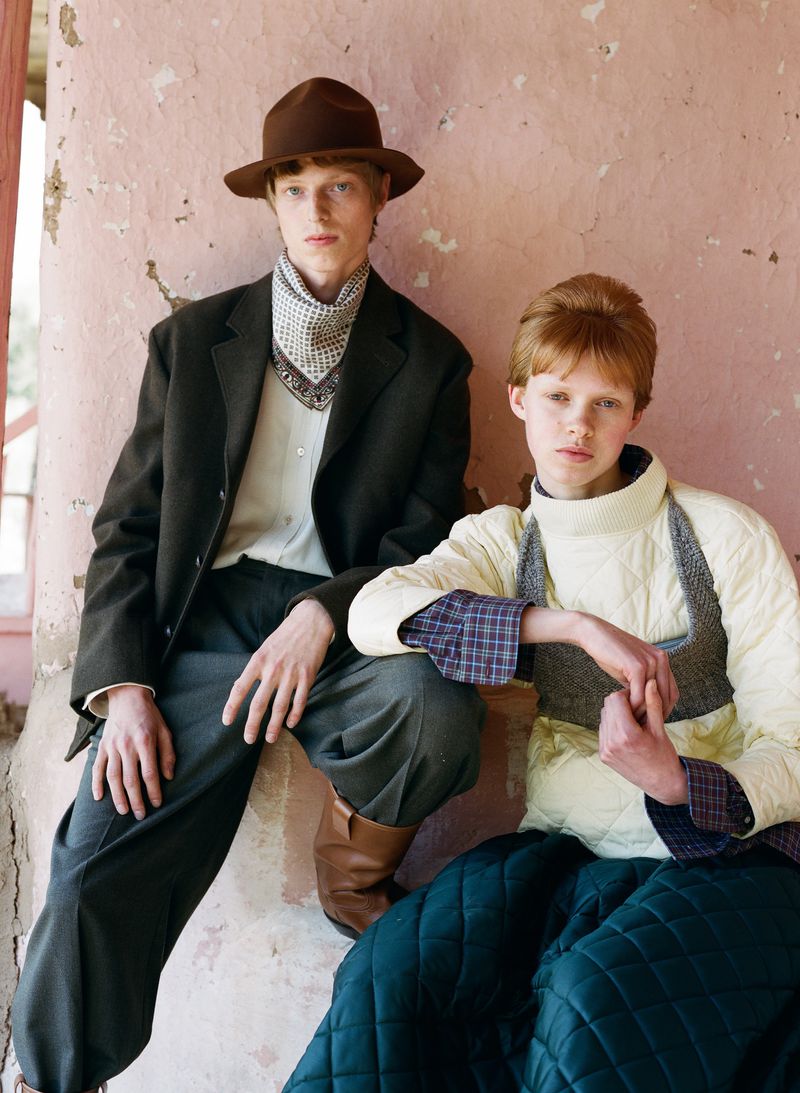 Two people in vintage-inspired fashion pose in front of a weathered wall. The models wear hats, vests, and quilted outerwear, set indoors.