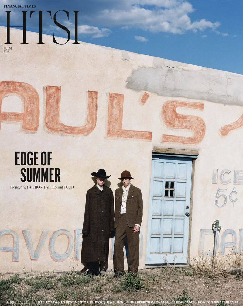 Two men pose in vintage suits and hats in front of a painted wall under a bright sky for a fashion editorial, capturing a summer edge.