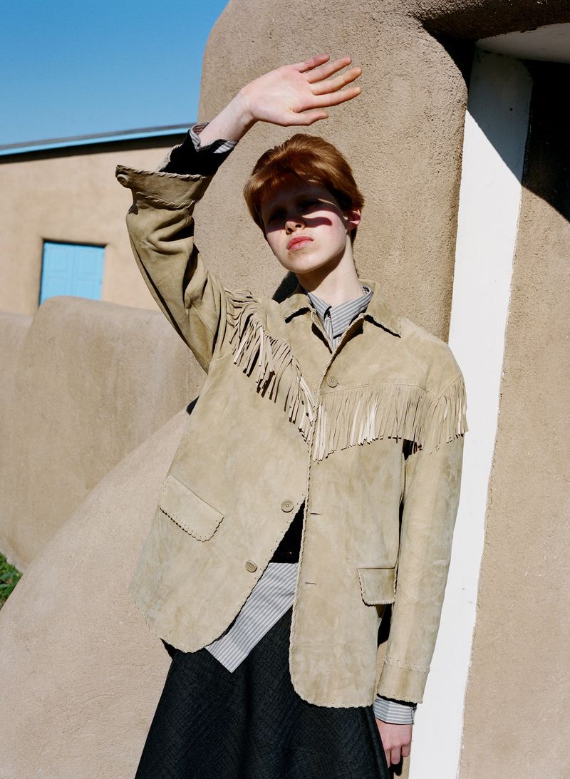 A stylish person in a fringe jacket stands in front of an adobe building, hand up to block the sunlight on a clear day outdoors.