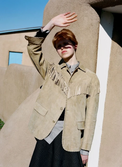 A stylish person in a fringe jacket stands in front of an adobe building, hand up to block the sunlight on a clear day outdoors.