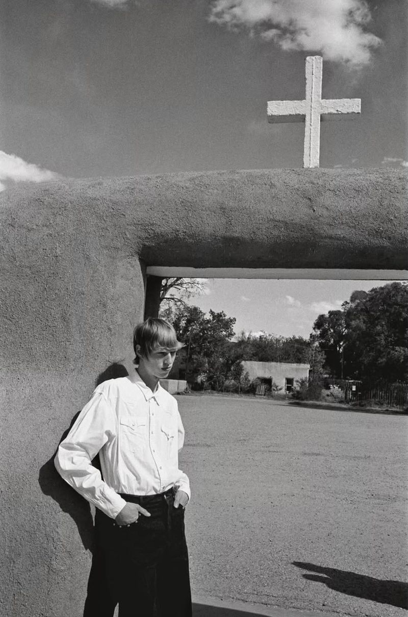 Black and white photo of a blonde man in a white shirt leaning against a stucco wall beneath a cross-topped archway outdoors.