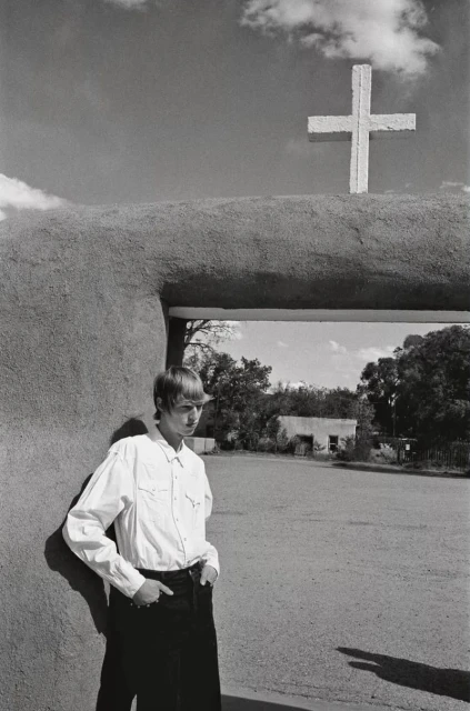 Black and white photo of a blonde man in a white shirt leaning against a stucco wall beneath a cross-topped archway outdoors.