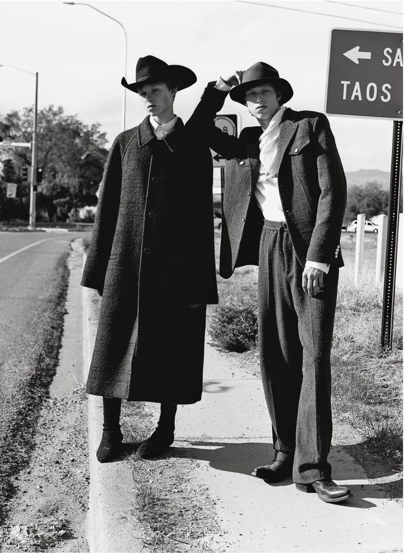Two adult men wearing dark colored hats and suits stand next to a road sign for Santa Fe and Taos on a rural roadside.