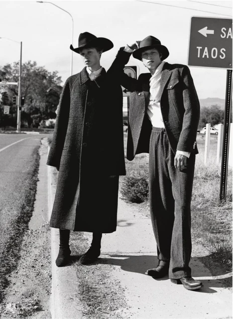 Two adult men wearing dark colored hats and suits stand next to a road sign for Santa Fe and Taos on a rural roadside.
