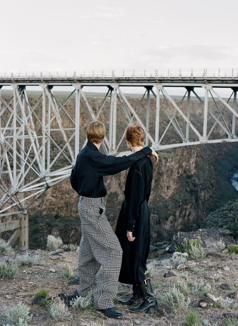 Two people stand near a bridge in black shirts and patterned pants in an outdoor setting with a rocky landscape and neutral sky.