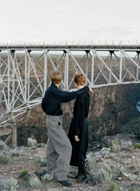 Two people stand near a bridge in black shirts and patterned pants in an outdoor setting with a rocky landscape and neutral sky.
