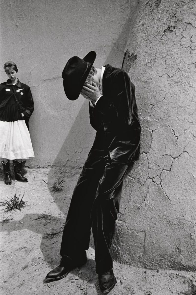 Black and white photograph of a man in a suit and cowboy hat leaning against a wall, with a girl in the background.