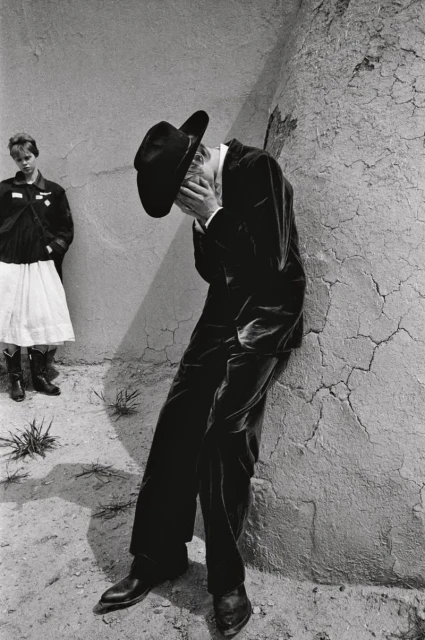 Black and white photograph of a man in a suit and cowboy hat leaning against a wall, with a girl in the background.
