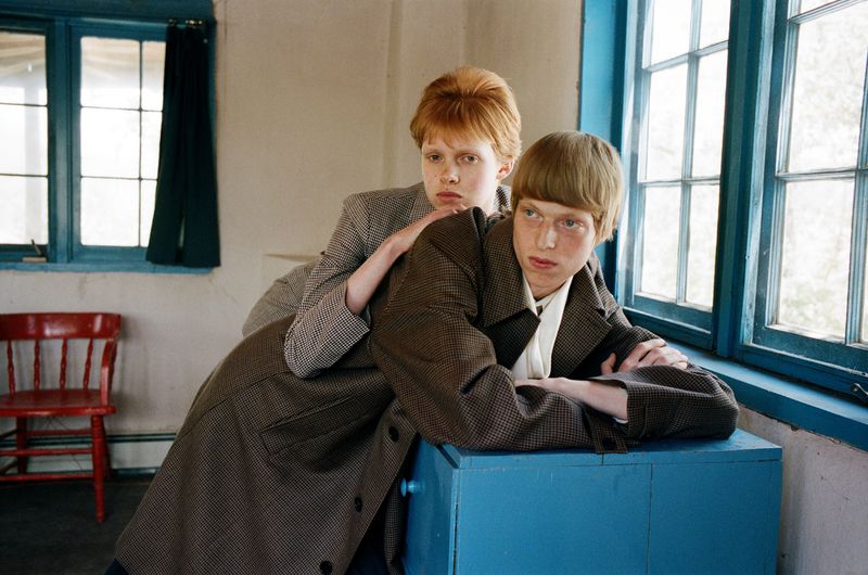 Two young adults pose in a room with a vintage aesthetic. One sits on a blue table while the other stands behind, showcasing retro fashion.