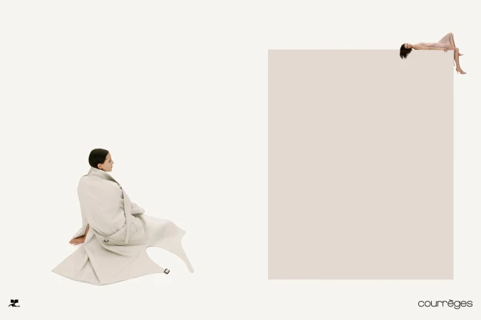 Model wrapped in a light beige coat seated on white background with a second model lying atop a large rectangular block