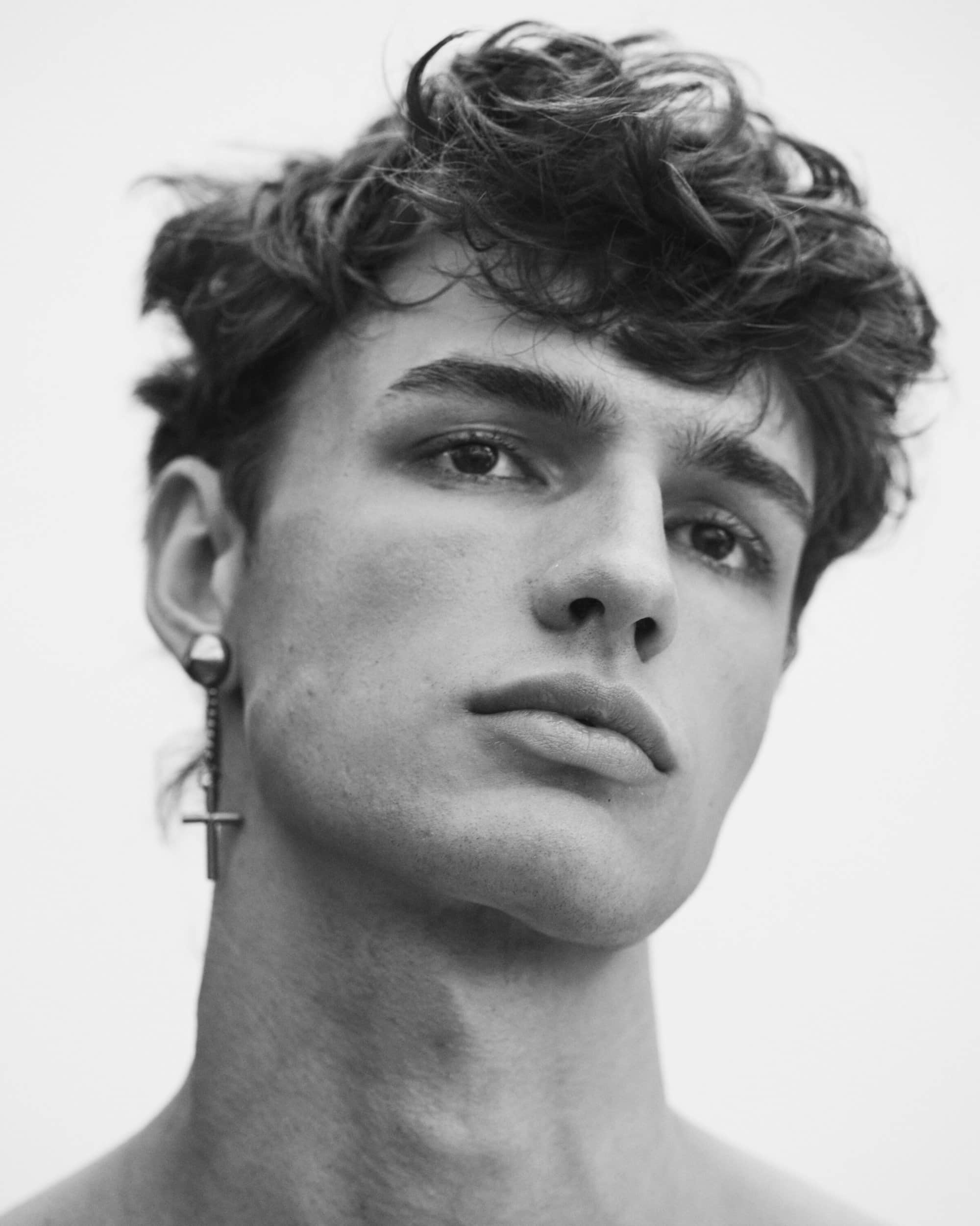 Close-up portrait of a male model with dark curly hair and an earring against a bright background for Dsquared2.