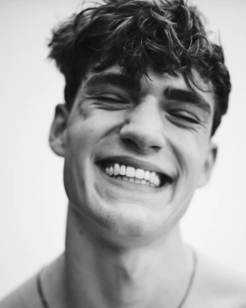 Close-up black and white portrait of a smiling young man with dark textured hair against a plain light background