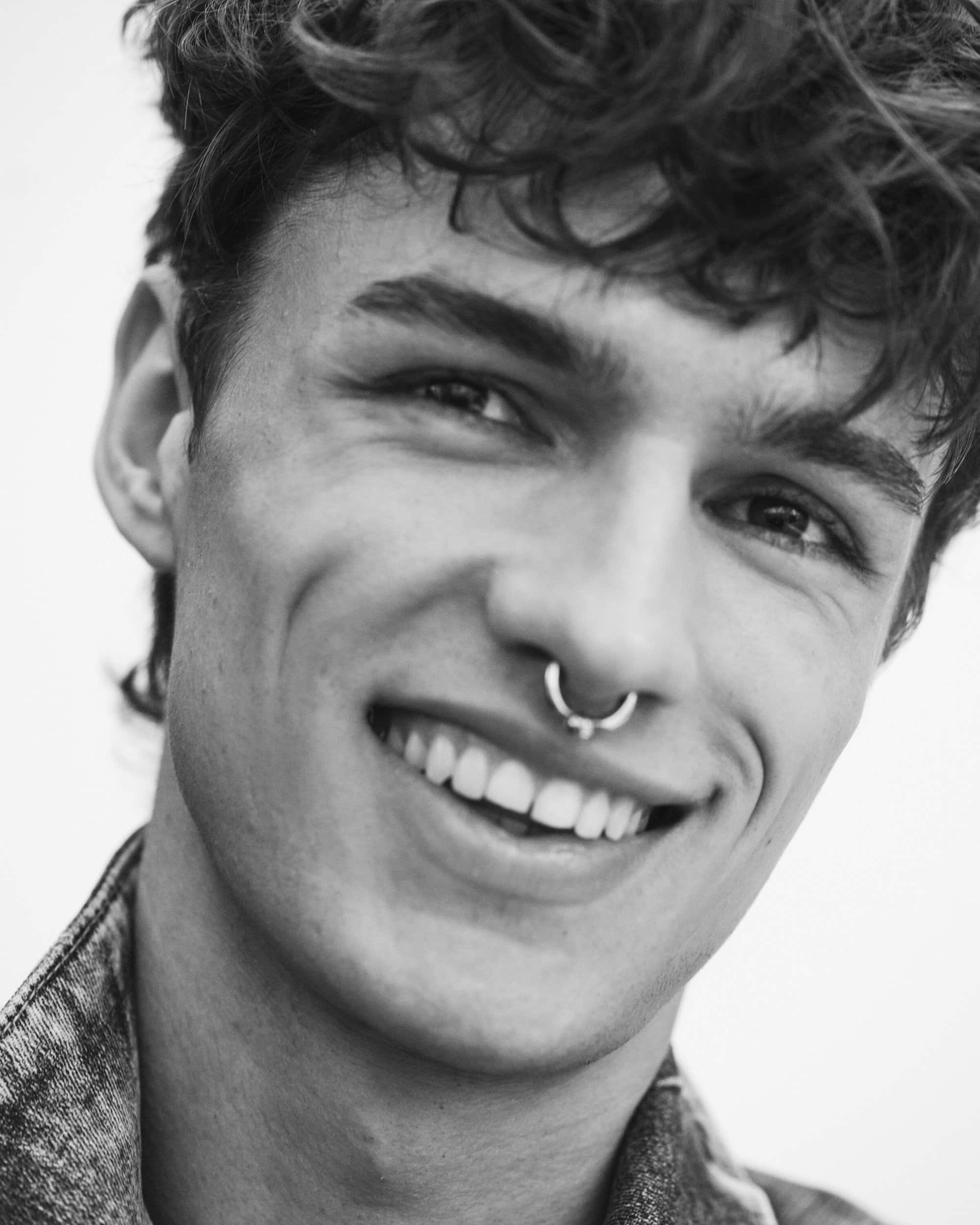 Close-up monochrome portrait of a smiling young man wearing a septum ring and denim collar against a white background