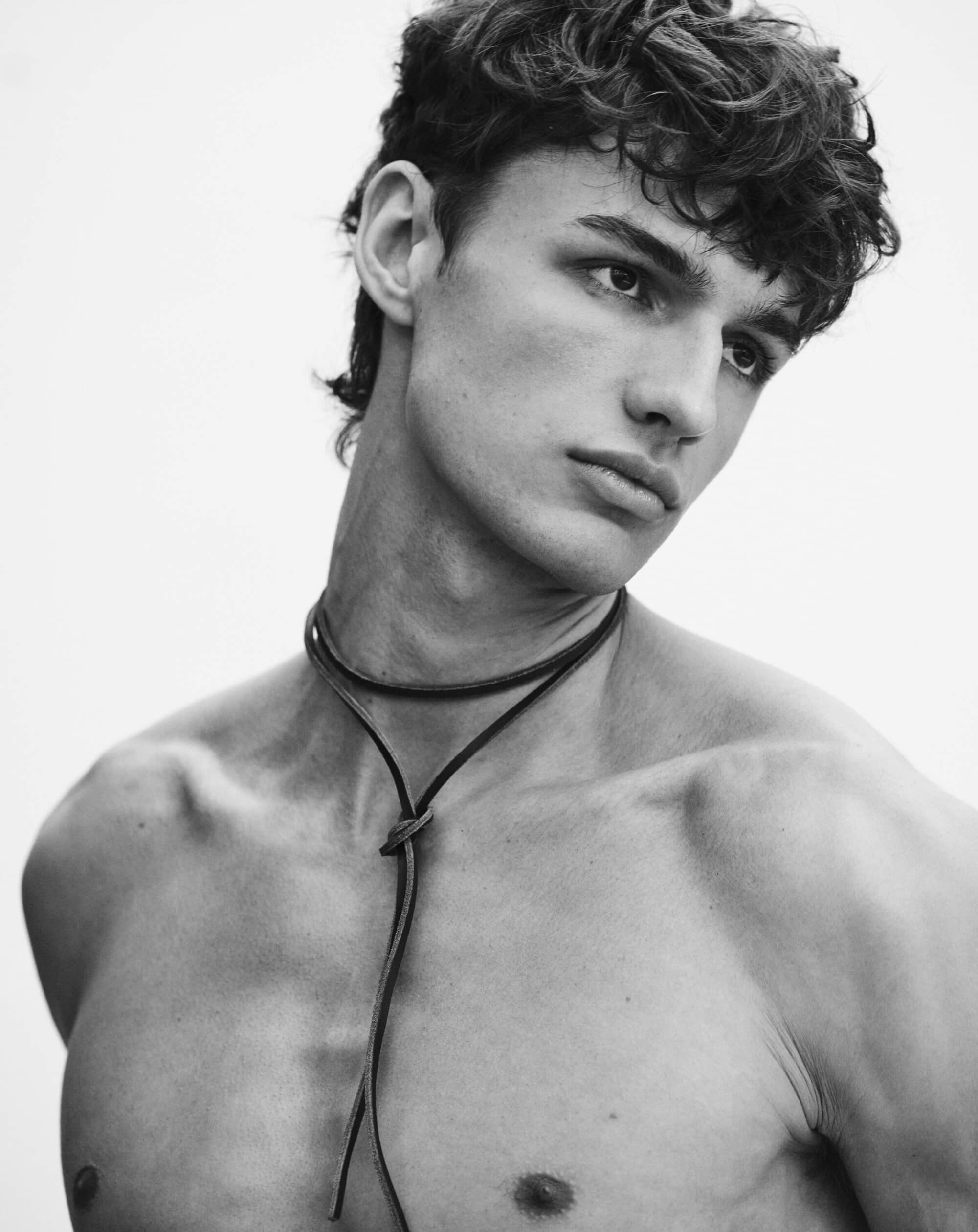 Portrait of a shirtless young male model wearing a thin leather cord necklace against a plain white background.