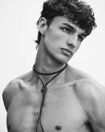 Portrait of a shirtless young male model wearing a thin leather cord necklace against a plain white background.