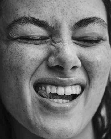 Close-up monochrome portrait of a laughing woman scrunching her nose with visible freckles and teeth