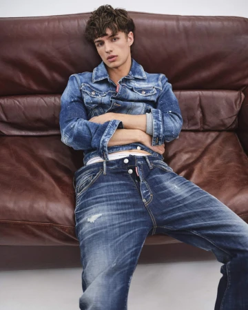 Young man wearing a blue denim jacket and jeans seated on a large brown leather couch for Dsquared2