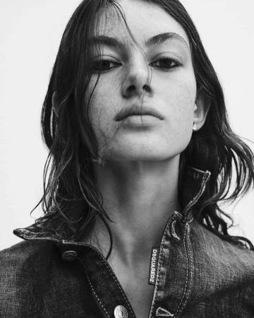 Close-up portrait of a model with dark tousled hair and freckles wearing a denim jacket against a white background.