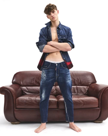 Young male model posing in denim shirt and jeans standing barefoot before a brown leather sofa