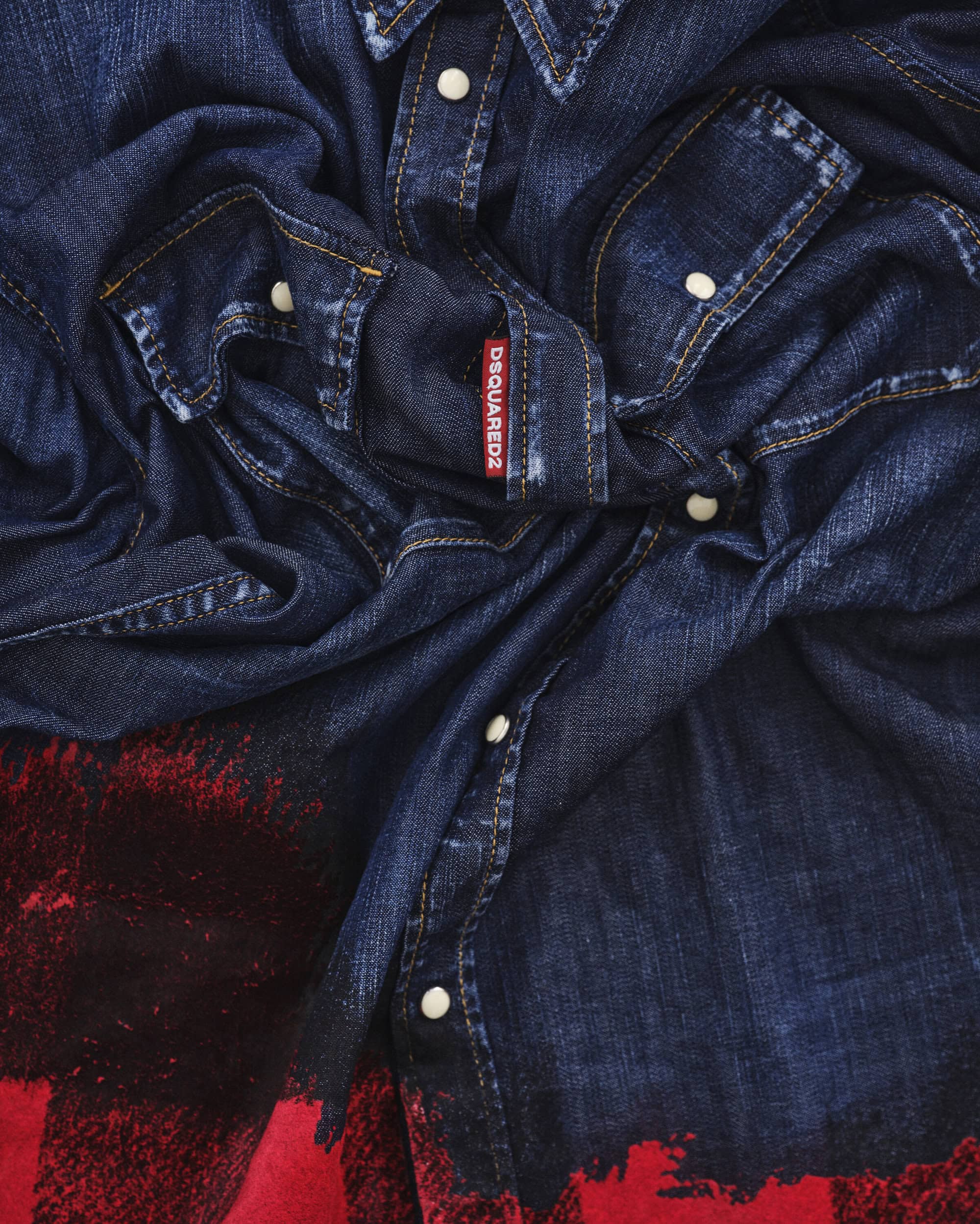 Close-up of a dark blue denim shirt with white snap buttons, contrast stitching, and a red brand label for the Dsquared2 campaign.