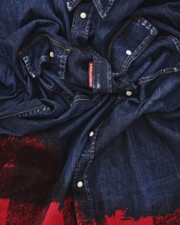 Close-up of a dark blue denim shirt with white snap buttons, contrast stitching, and a red brand label for the Dsquared2 campaign.