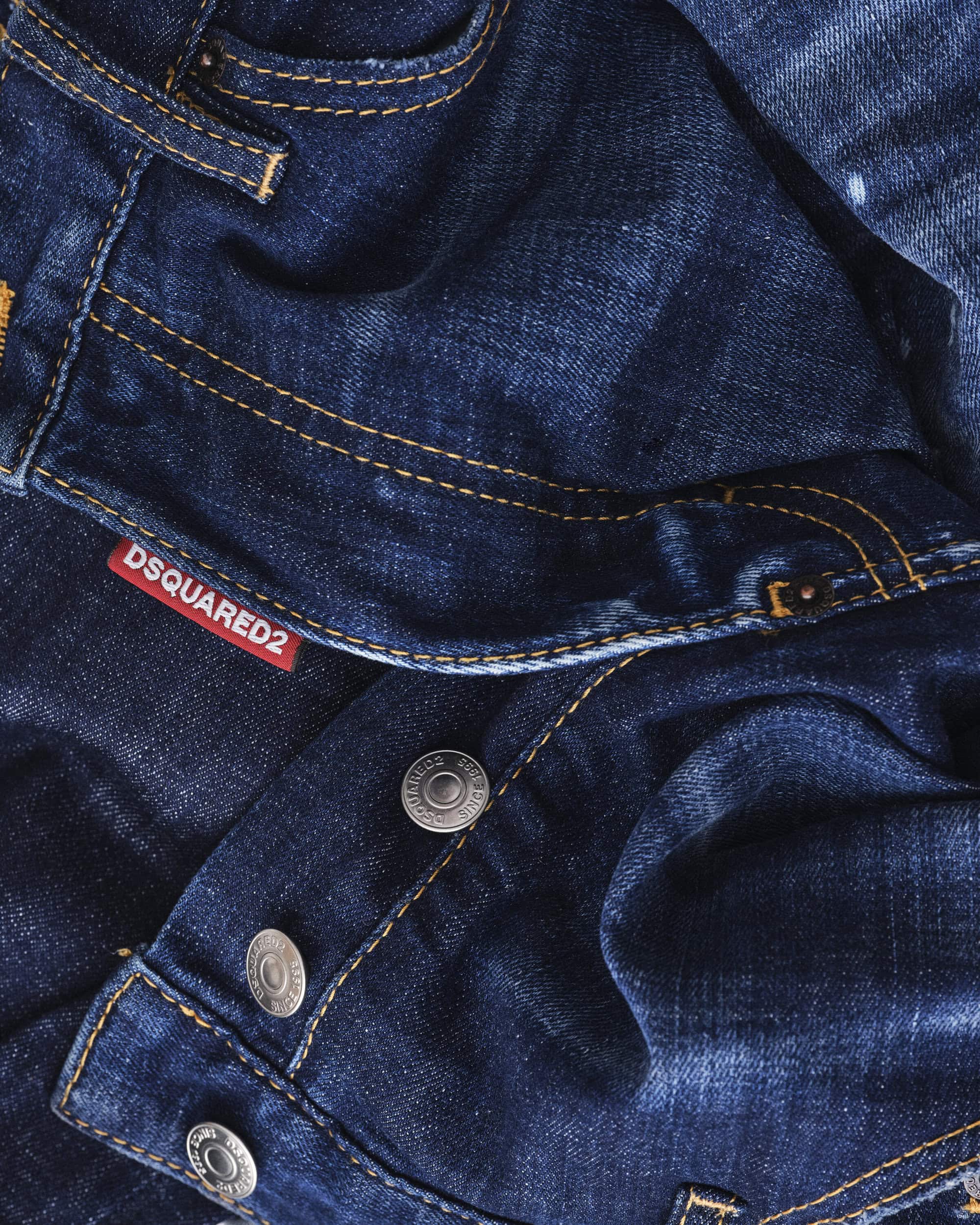 Close-up macro shot of dark blue denim jeans with orange topstitching, metallic buttons, and a red Dsquared2 label