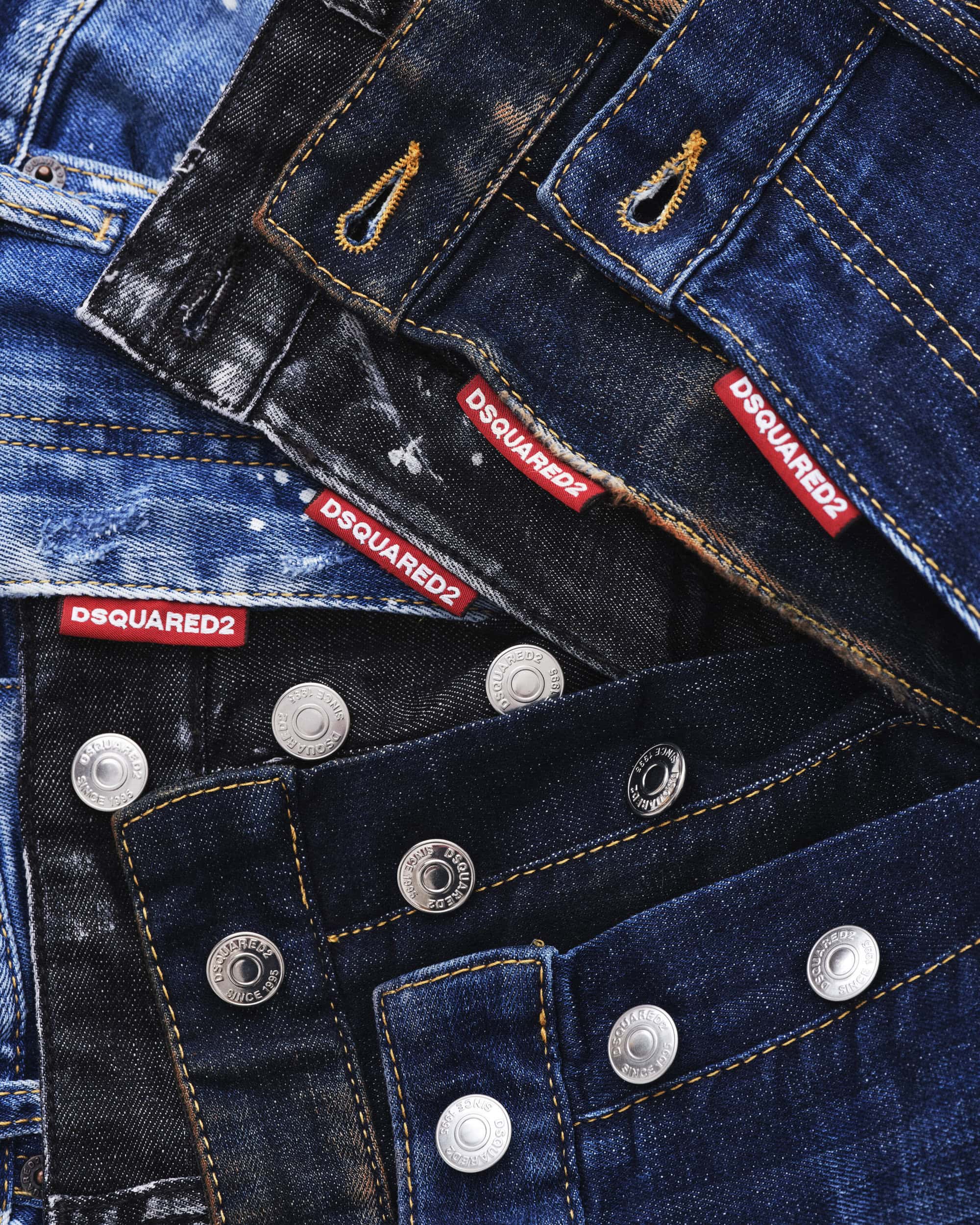 Stacked layers of blue and dark denim jeans featuring metallic buttons and red DSQUARED2 branded tags.