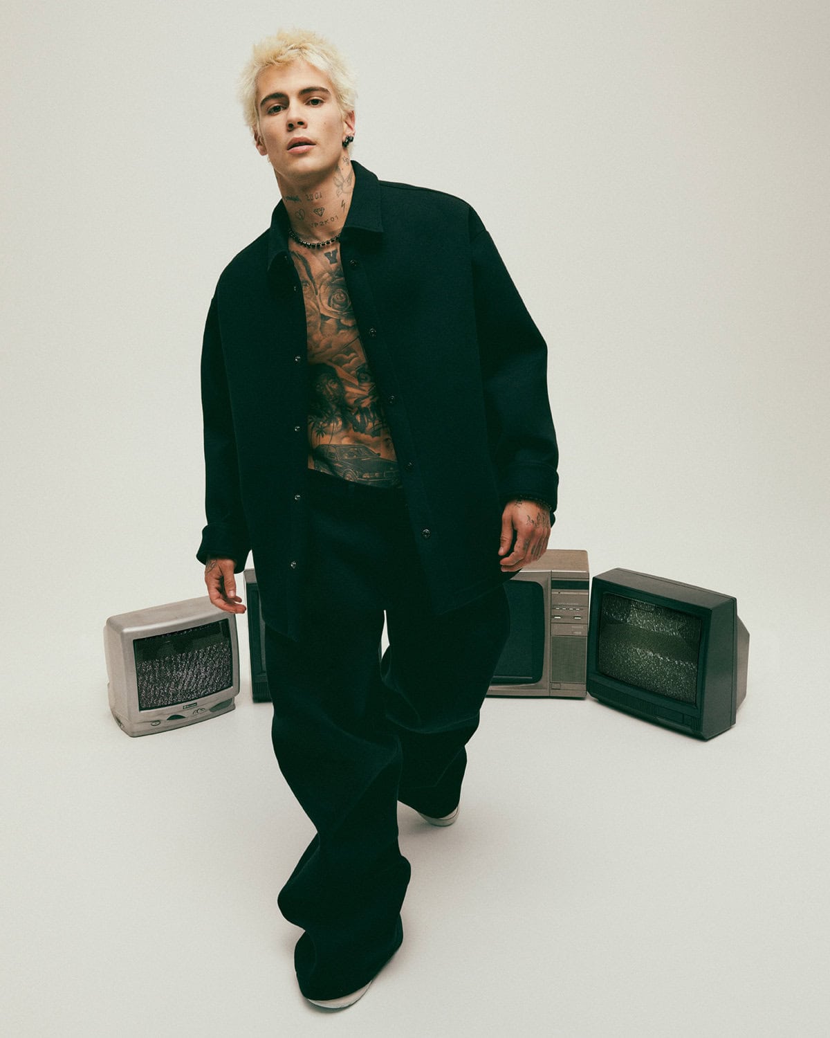 Blonde male model with neck and chest tattoos wearing a dark shirt against a seamless white background with vintage televisions