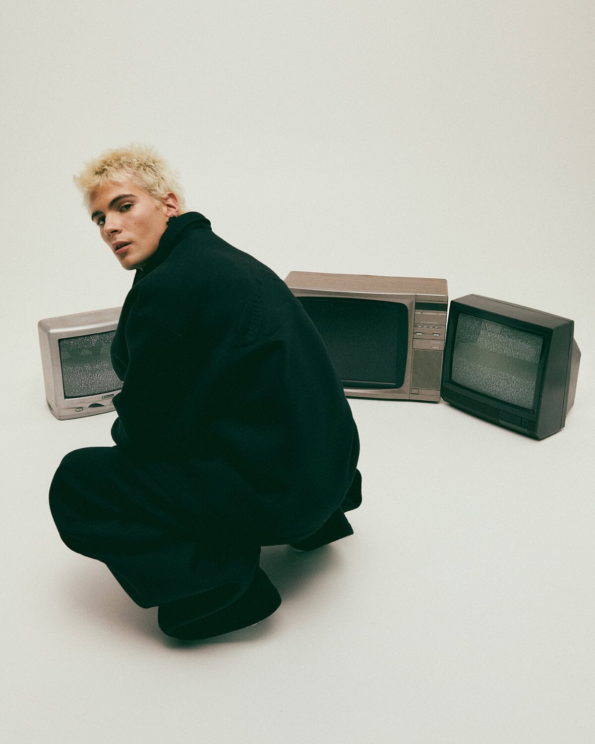 Blonde male model wearing a dark coat crouching amidst vintage CRT televisions against a white background.
