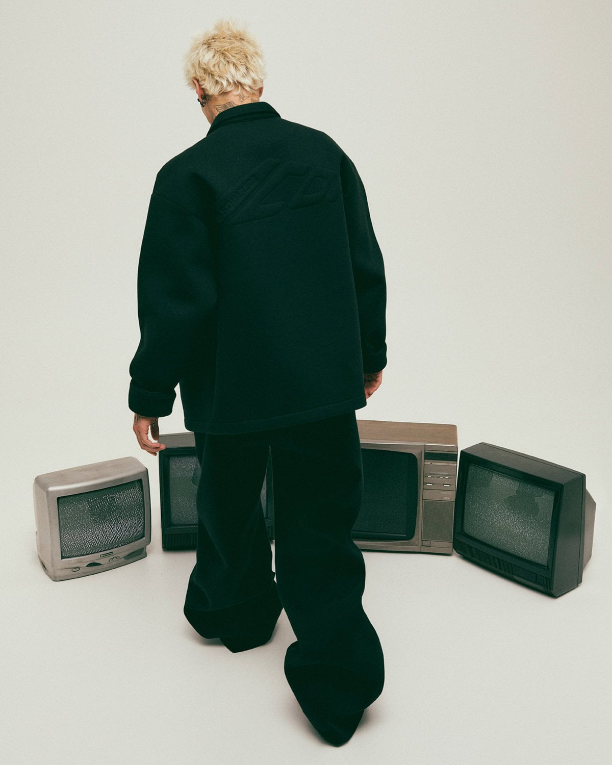 Model seen from behind wearing a black jacket and matching trousers surrounded by four vintage cathode ray tube televisions.