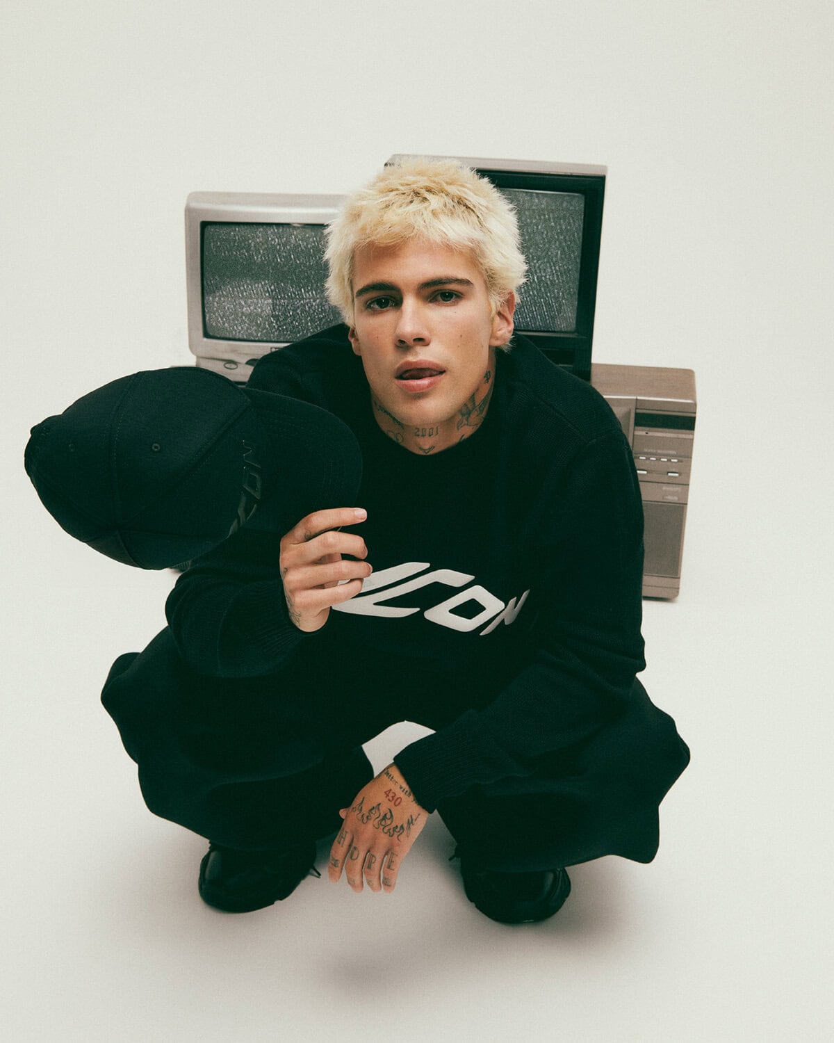 Blonde male model squatting while holding a black cap in front of vintage televisions for Dsquared2
