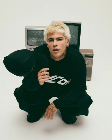 Blonde male model squatting while holding a black cap in front of vintage televisions for Dsquared2