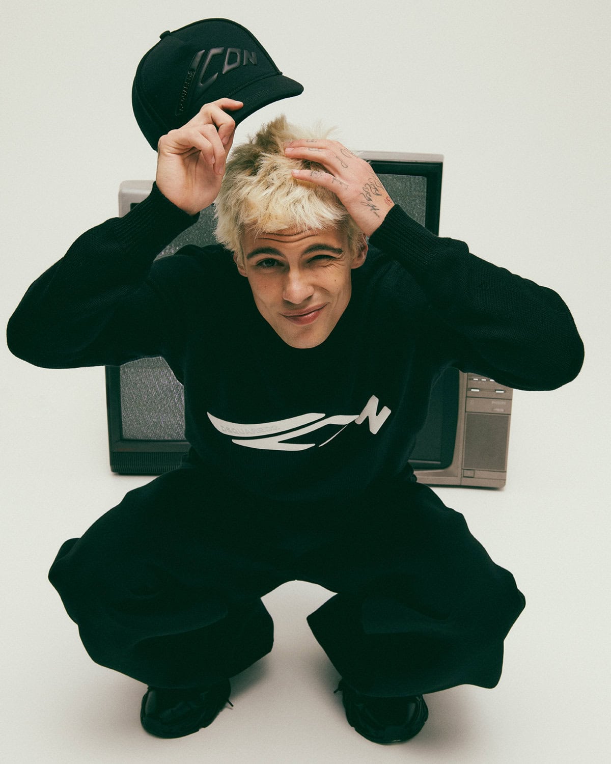 Blonde male model wearing black sweater and holding baseball cap in front of vintage televisions