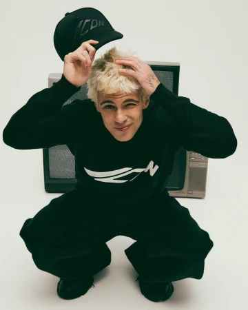Blonde male model wearing black sweater and holding baseball cap in front of vintage televisions