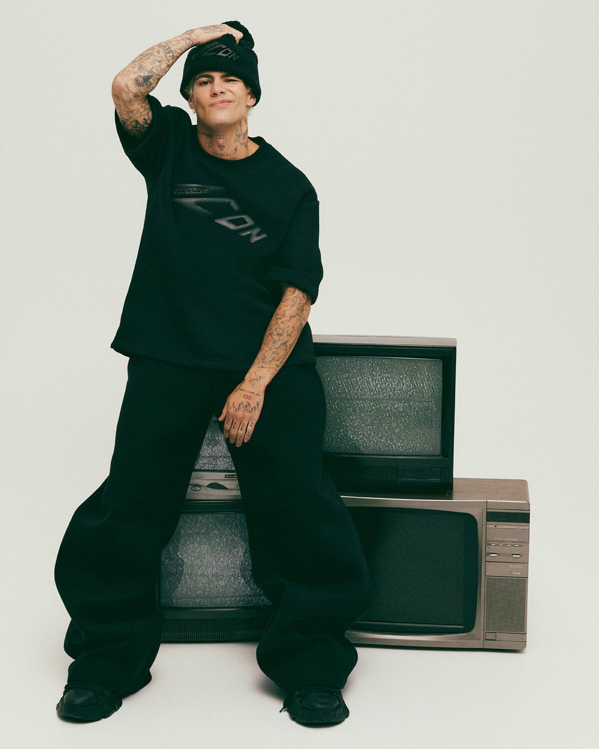 Man wearing a black t-shirt, beanie, and trousers posing against a stack of vintage televisions in a studio setting.