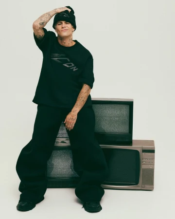 Man wearing a black t-shirt, beanie, and trousers posing against a stack of vintage televisions in a studio setting.