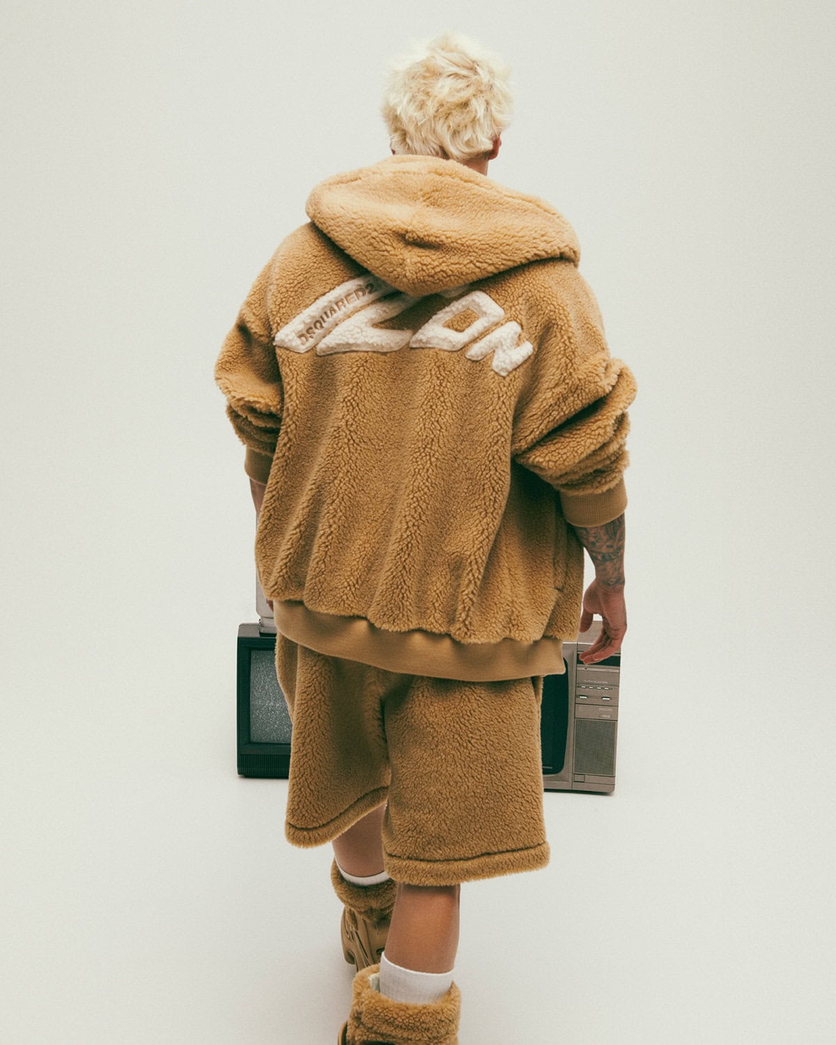 Model with blonde hair wearing tan fleece hoodie and shorts facing away from camera against a white backdrop