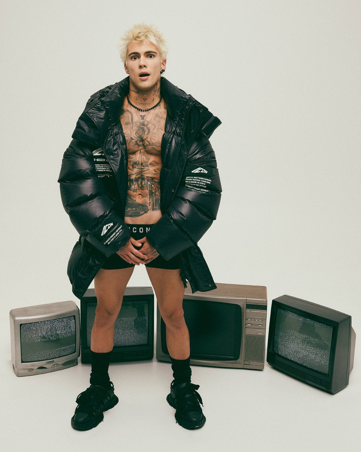 Blonde male model wearing an oversized black puffer jacket, black underwear, and black sneakers surrounded by old televisions against a white background.
