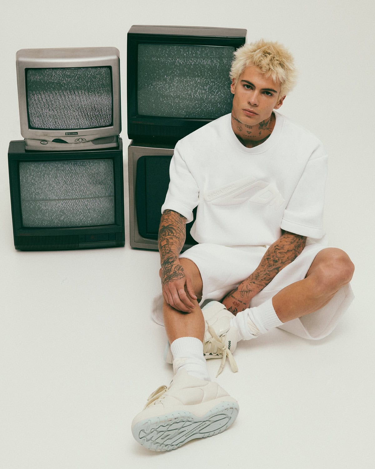 Blonde male model wearing white sweatshirt and shorts sitting next to stacked vintage televisions on a white background