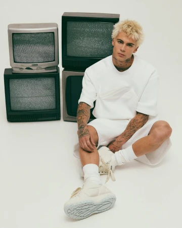 Blonde male model wearing white sweatshirt and shorts sitting next to stacked vintage televisions on a white background