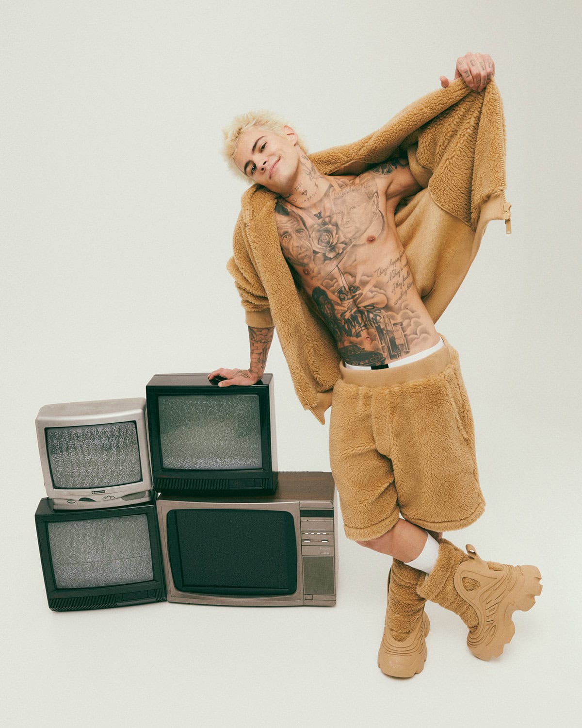 Man wearing a camel fleece hoodie and shorts leaning against stacked vintage televisions against a plain white background.