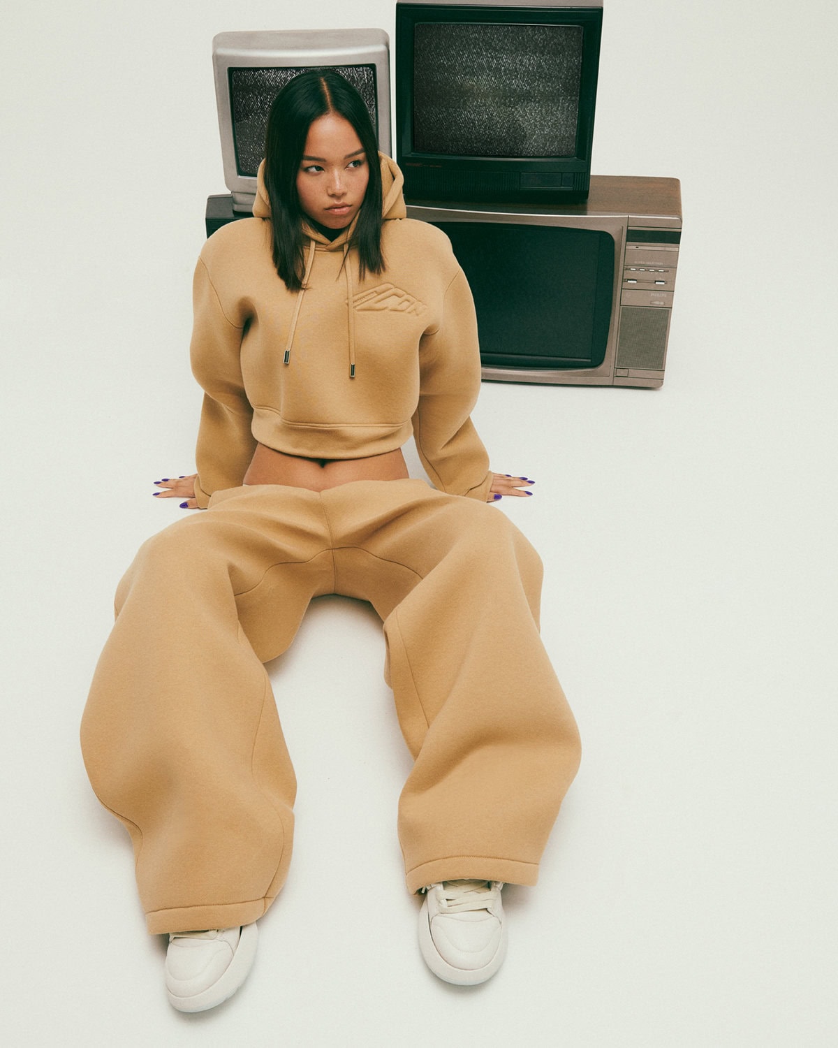 Model wearing a camel hoodie and matching sweatpants seated against a white seamless background with stacked vintage televisions.