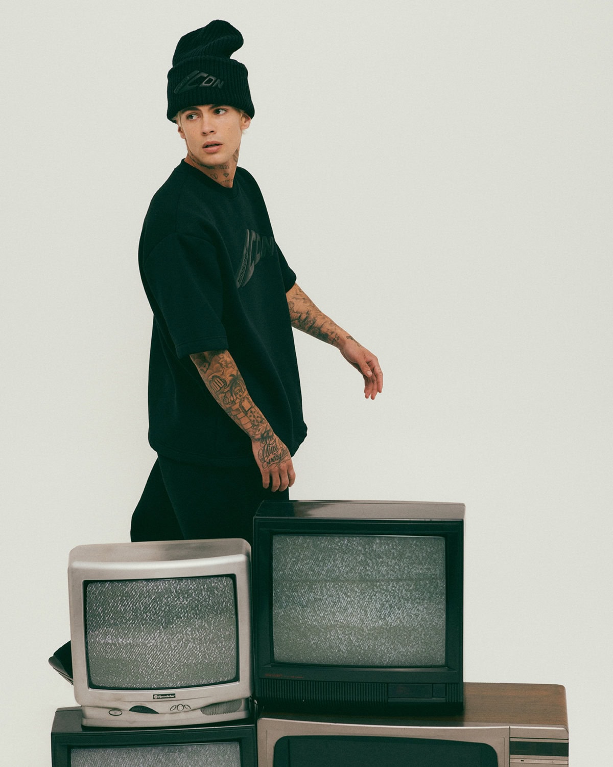 Young man wearing black t-shirt and beanie standing near stacked vintage television sets against a white background.