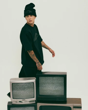 Young man wearing black t-shirt and beanie standing near stacked vintage television sets against a white background.