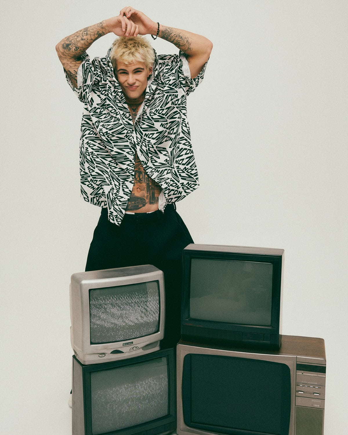 Blonde male model with tattoos posing behind a stack of vintage CRT television sets against a plain background