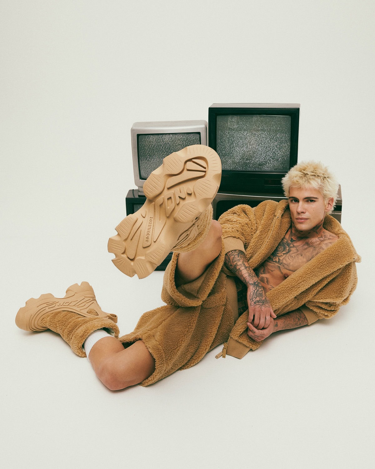 Man wearing a camel sherpa fleece robe and matching chunky sneakers lying against vintage televisions on a white background.