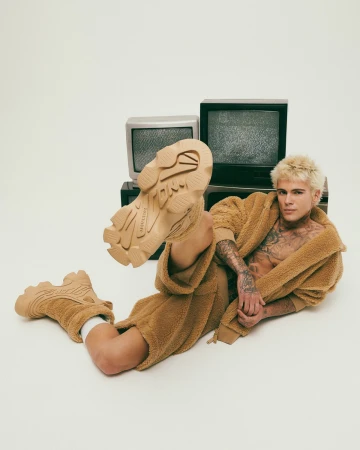 Man wearing a camel sherpa fleece robe and matching chunky sneakers lying against vintage televisions on a white background.