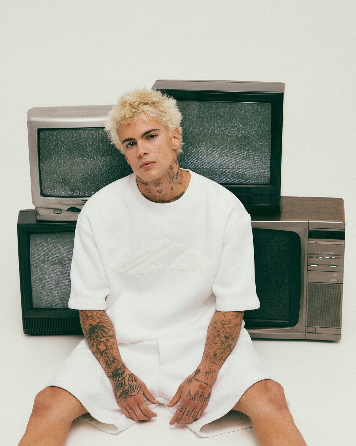 Young man with bleached hair and tattoos wearing a white t-shirt sitting amidst a stack of vintage television sets