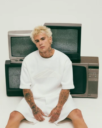 Young man with bleached hair and tattoos wearing a white t-shirt sitting amidst a stack of vintage television sets